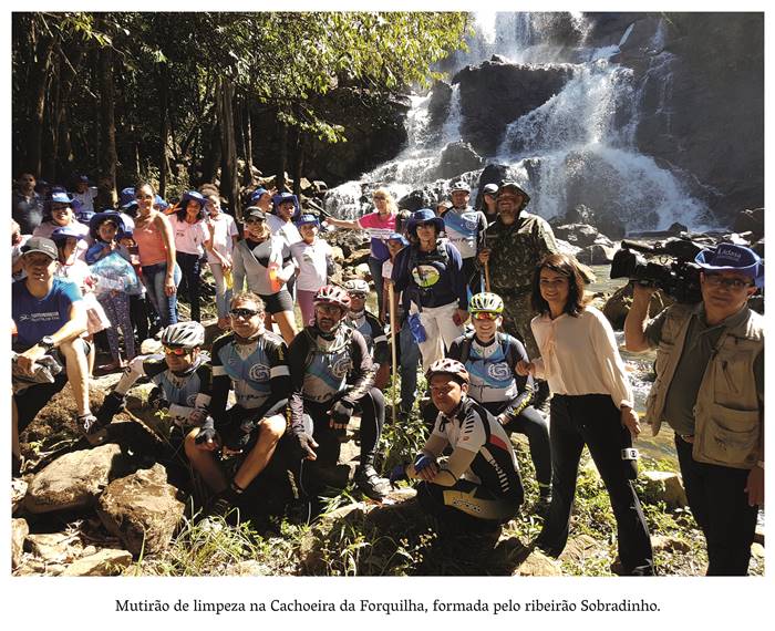  4 u- Mutirão de limpeza da Cachoeira da Forquilha, composta pelo Ribeirão Sobradinho. Um grupo de homens e mulheres estão reunidos posando para uma fotografia,. O local está iluminado pelo sol e ao fundo uma cachoeira com água límpida e cristalina, que, encontra-se rodeada por arvoredos. Realização: Academia Planaltinense de Letras, Artes e Ciências (APLAC), pelo Ecomuseu Pedra Fundamental e pelo Coletivo Nativo Audiodescrição produzida  pelo Instituto de Promoção das Pessoas com Deficiência Visual Audiodescritora: Elma Lúcia Rodrigues Consultor: Fernando Rodrigues Este projeto é promovido com recursos do Fundo de Apoio a Cultura do DF